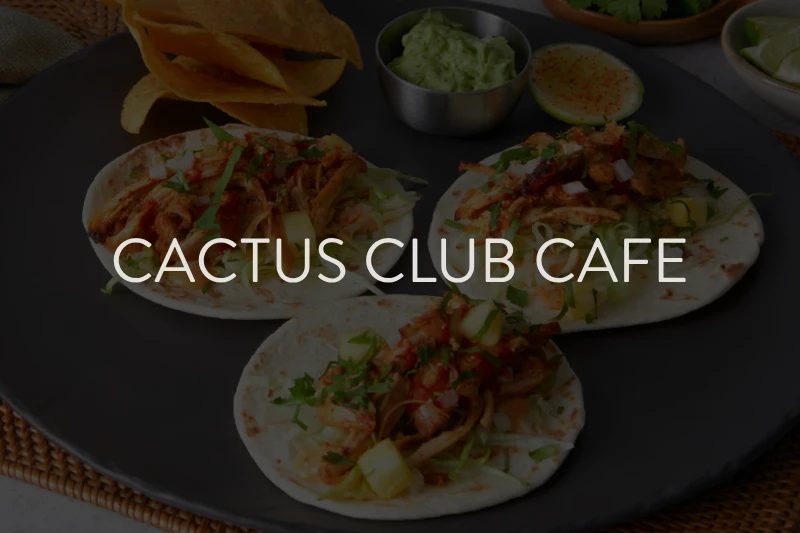 Three pork tacos with toppings on a white plate beside chips and guacamole, with the text "Cactus Club Cafe" overlaid.